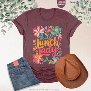 Pode incluir: Uma camiseta bord&ocirc; com a frase "Lunch Lady" rodeada por desenhos florais coloridos. A imagem tamb&eacute;m inclui um par de jeans azuis e um chap&eacute;u de cowboy marrom.