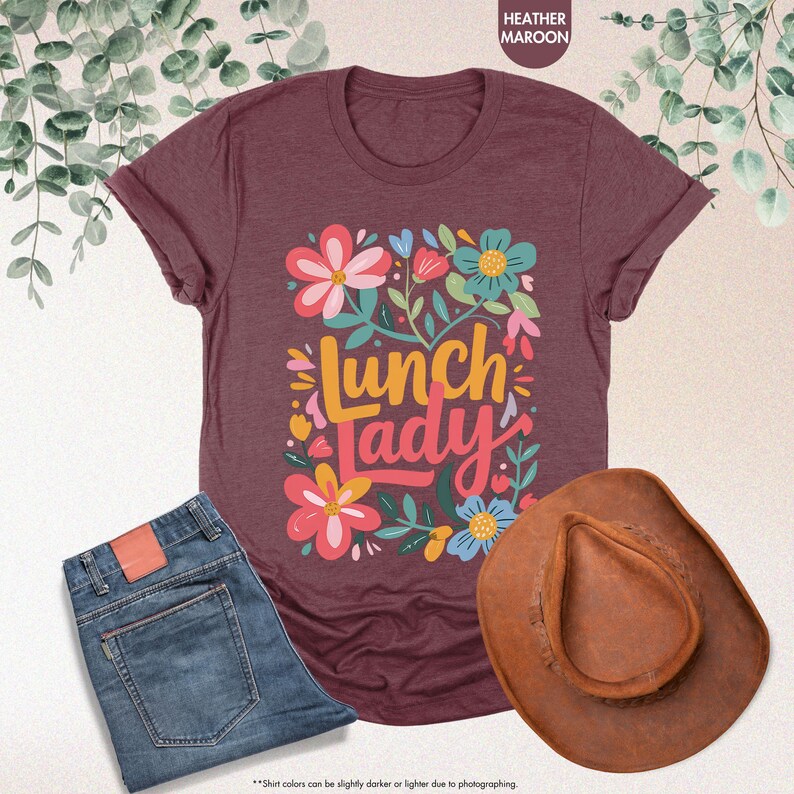 Pode incluir: Uma camiseta bord&ocirc; com a frase "Lunch Lady" rodeada por desenhos florais coloridos. A imagem tamb&eacute;m inclui um par de jeans azuis e um chap&eacute;u de cowboy marrom.