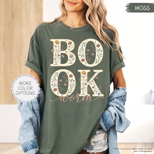 以下が含まれることがあります： モスグリーンのTシャツ。白い文字で「BOOK WORM」と書かれ、花柄のデザインが施されています。文字は白で、様々な色の花のアクセントがあります。シャツは袖をまくり、ゆったりとしたフィット感です。「MORE COLOR OPTIONS」という文字が白い円の中にあります。