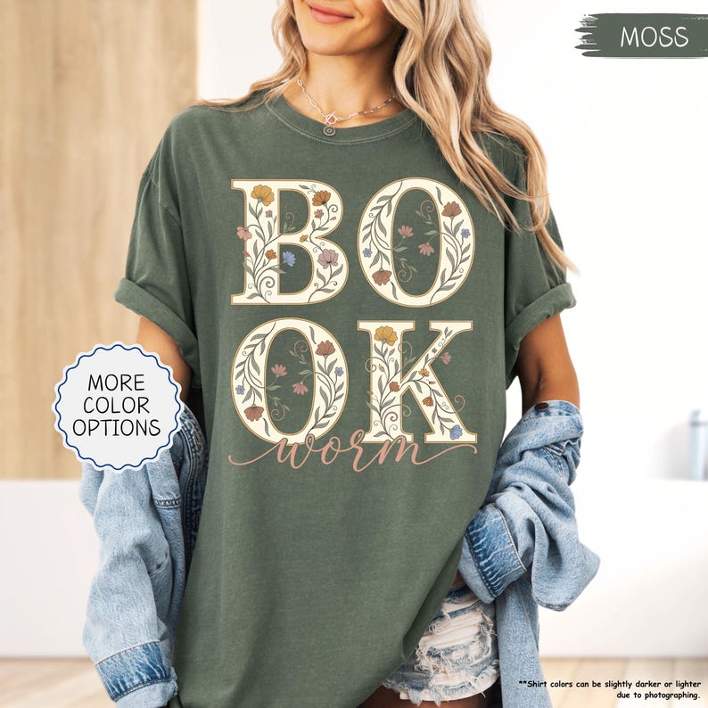 以下が含まれることがあります： モスグリーンのTシャツ。白い文字で「BOOK WORM」と書かれ、花柄のデザインが施されています。文字は白で、様々な色の花のアクセントがあります。シャツは袖をまくり、ゆったりとしたフィット感です。「MORE COLOR OPTIONS」という文字が白い円の中にあります。