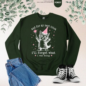 May include: A forest green long-sleeve sweatshirt featuring a white graphic of a kitten wearing a pink party hat and holding a star wand. The text reads, "and for my next trick i'll forget what i was doing," surrounded by small pink stars.