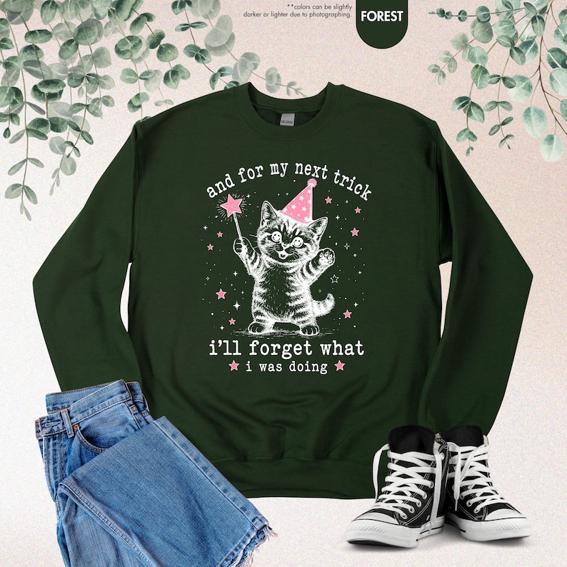 May include: A forest green long-sleeve sweatshirt featuring a white graphic of a kitten wearing a pink party hat and holding a star wand. The text reads, "and for my next trick i'll forget what i was doing," surrounded by small pink stars.