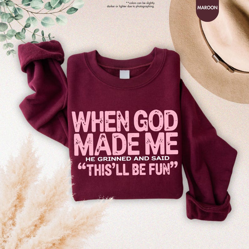 May include: Maroon sweatshirt with the text "WHEN GOD MADE ME HE GRINNED AND SAID 'THIS'LL BE FUN'" in a light pink font. The sweatshirt has a crew neck and long sleeves. The word "MAROON" is in a circle in the upper right corner.