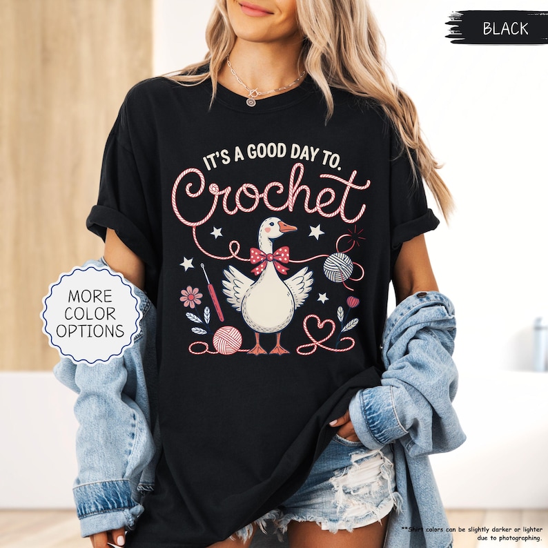 May include: Black t-shirt with the text "IT'S A GOOD DAY TO. Crochet" in a whimsical font. The graphic features a goose wearing a bow tie, surrounded by yarn balls, flowers, and crochet hooks. The words "MORE COLOR OPTIONS" are also visible.