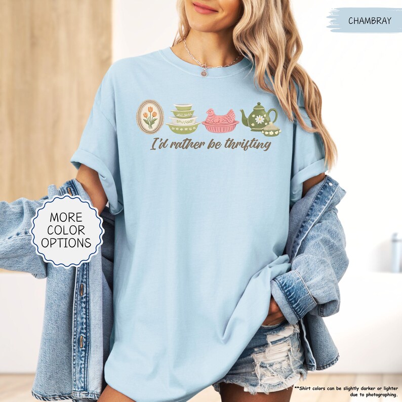 Pu&ograve; includere: T-shirt azzurra con la scritta "I'd rather be thrifting" e una grafica di utensili da cucina vintage. La maglietta ha un girocollo e maniche corte. Le parole "More Color Options" sono in un cerchio bianco.