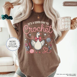 May include: Espresso-colored t-shirt with the text "It's a good day to Crochet" in a whimsical font. The graphic features a goose wearing a bow tie, surrounded by yarn balls, flowers, and crochet hooks. The shirt also has "More Color Options" text.
