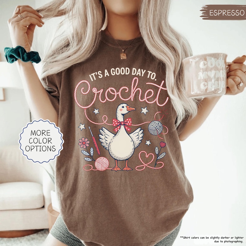 May include: Espresso-colored t-shirt with the text "It's a good day to Crochet" in a whimsical font. The graphic features a goose wearing a bow tie, surrounded by yarn balls, flowers, and crochet hooks. The shirt also has "More Color Options" text.