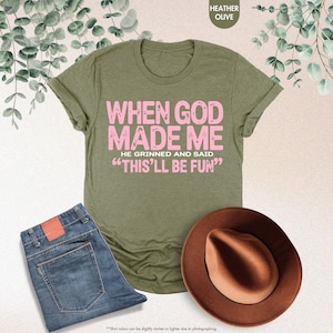 May include: An olive green t-shirt with the text "WHEN GOD MADE ME HE GRINNED AND SAID 'THIS'LL BE FUN'" in pink. The image also shows a pair of blue jeans and a brown hat. The shirt's color is described as Heather Olive.