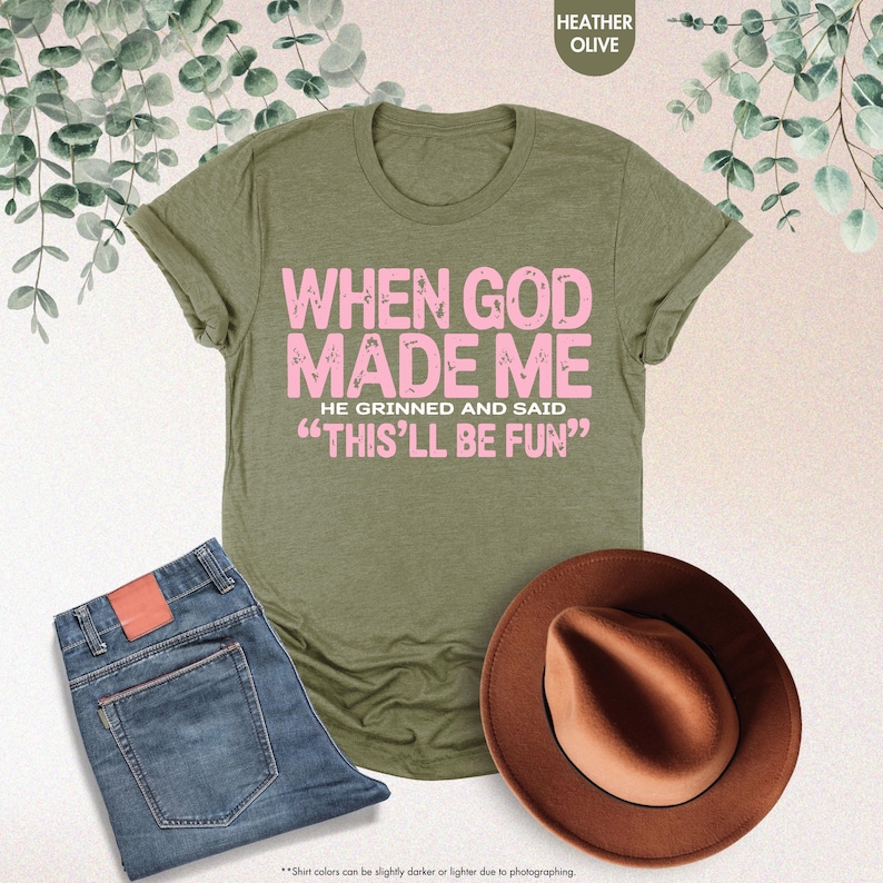 May include: An olive green t-shirt with the text "WHEN GOD MADE ME HE GRINNED AND SAID 'THIS'LL BE FUN'" in pink. The image also shows a pair of blue jeans and a brown hat. The shirt's color is described as Heather Olive.