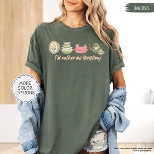 Pu&ograve; includere: T-shirt verde muschio con la scritta "I'd rather be thrifting" e una grafica di utensili da cucina vintage. La grafica include un design floreale incorniciato, ciotole, un pollo rosa e una teiera. La maglietta ha le maniche arrotolate.