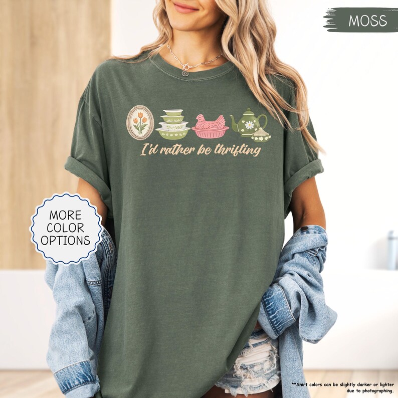 Pu&ograve; includere: T-shirt verde muschio con la scritta "I'd rather be thrifting" e una grafica di utensili da cucina vintage. La grafica include un design floreale incorniciato, ciotole, un pollo rosa e una teiera. La maglietta ha le maniche arrotolate.