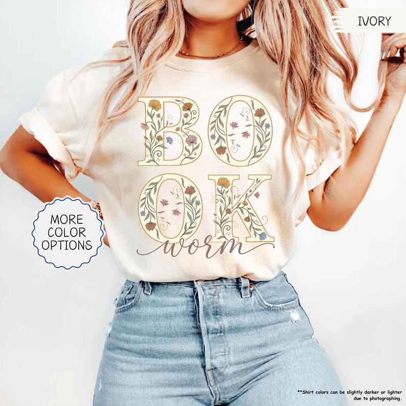 以下が含まれることがあります： フローラルデザインの「BOOK worm OK」という言葉が入ったアイボリー色のTシャツ。文字は茶色、青、紫の色合いの花とつるで埋められています。シャツには「More Color Options」ボタンもあります。