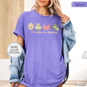 Pu&ograve; includere: Una maglietta viola con la scritta "I'd rather be thrifting" e illustrazioni di un fiore incorniciato, piatti, un pollo e una teiera. La maglietta ha le maniche arrotolate. &Egrave; visibile anche il testo "MORE COLOR OPTIONS".