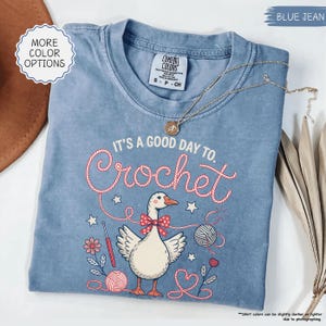 May include: A blue jean Comfort Colors t-shirt with the text "It's a good day to Crochet" and a graphic of a goose wearing a bow tie. The shirt also features a gold necklace. The shirt is a size small.