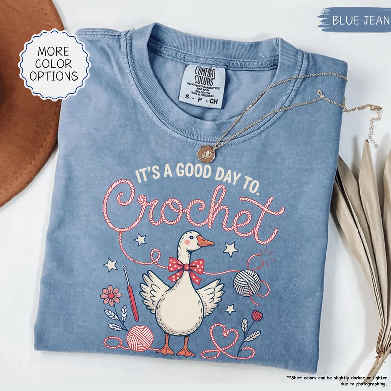 May include: A blue jean Comfort Colors t-shirt with the text "It's a good day to Crochet" and a graphic of a goose wearing a bow tie. The shirt also features a gold necklace. The shirt is a size small.