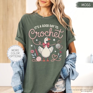 May include: Moss green t-shirt with the text "IT'S A GOOD DAY TO. Crochet" and a graphic of a goose wearing a bow tie, surrounded by crochet-related elements. The shirt has rolled-up sleeves and a relaxed fit. "MORE COLOR OPTIONS" is also visible.