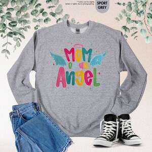 May include: A gray sweatshirt with the text "Mom of an Angel" in colorful letters and angel wings. The sweatshirt is paired with blue jeans and black and white sneakers, creating a casual, everyday look.