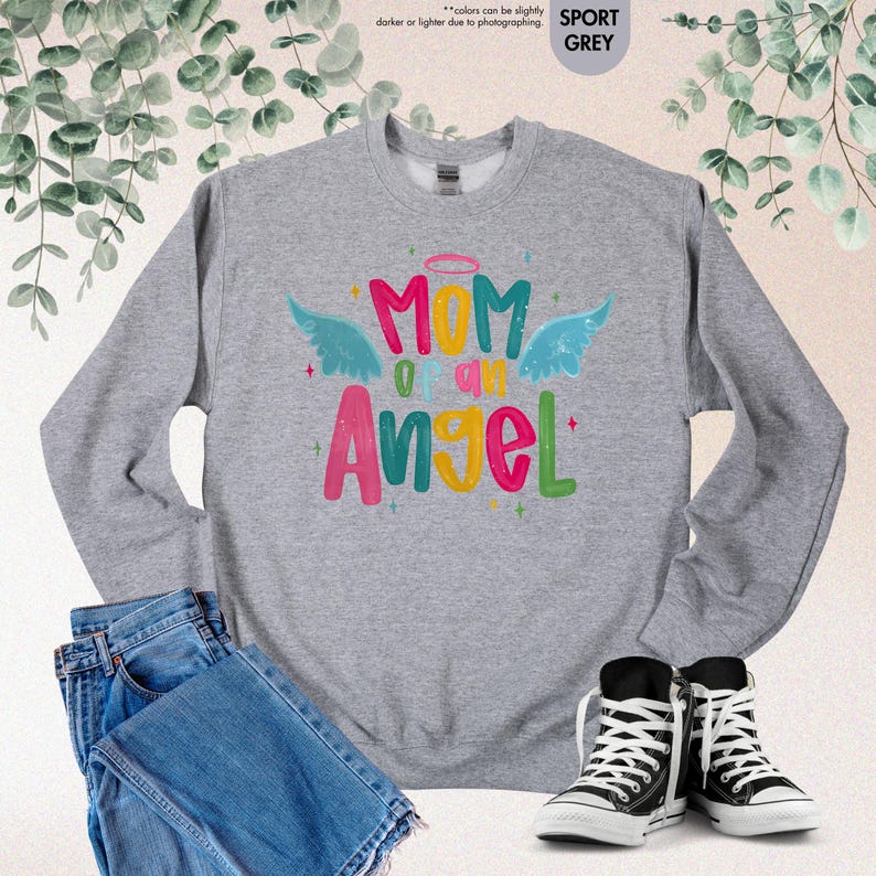 May include: A gray sweatshirt with the text "Mom of an Angel" in colorful letters and angel wings. The sweatshirt is paired with blue jeans and black and white sneakers, creating a casual, everyday look.