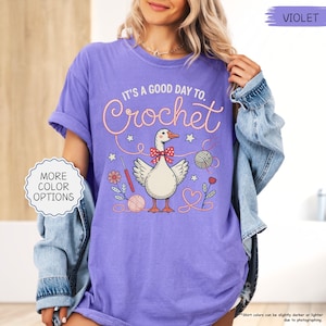 May include: A violet t-shirt with the text "IT'S A GOOD DAY TO. Crochet" in a whimsical font. The graphic features a goose with a bow tie, yarn, and crochet hooks. The shirt also has the text "MORE COLOR OPTIONS".