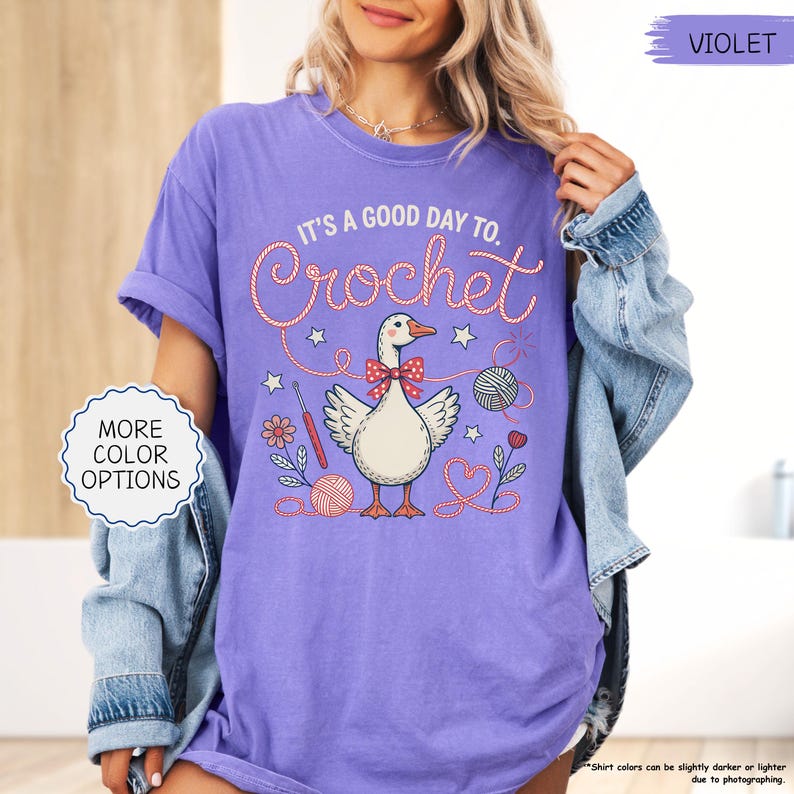 May include: A violet t-shirt with the text "IT'S A GOOD DAY TO. Crochet" in a whimsical font. The graphic features a goose with a bow tie, yarn, and crochet hooks. The shirt also has the text "MORE COLOR OPTIONS".