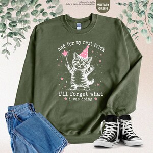 May include: A military green crewneck sweatshirt featuring a white graphic of a kitten wearing a pink party hat and holding a magic wand. The text reads, "and for my next trick i'll forget what i was doing," surrounded by small pink stars.