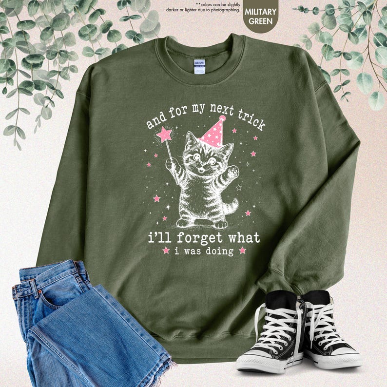 May include: A military green crewneck sweatshirt featuring a white graphic of a kitten wearing a pink party hat and holding a magic wand. The text reads, "and for my next trick i'll forget what i was doing," surrounded by small pink stars.