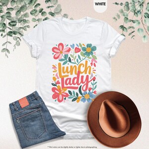 Pode incluir: Camiseta branca com a frase "Lunch Lady" em um design floral colorido. A imagem tamb&eacute;m mostra jeans azuis e um chap&eacute;u fedora marrom. A camiseta &eacute; branca.