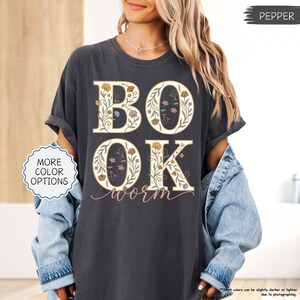 以下が含まれることがあります： 大きな花柄の文字で「BOOK」と「OK」と書かれた濃い灰色のTシャツ。 「worm」の文字が筆記体で下に書かれています。 シャツは袖をまくり、デニムジャケットを着ています。 「MORE COLOR OPTIONS」という文字が白い円の中にあります。