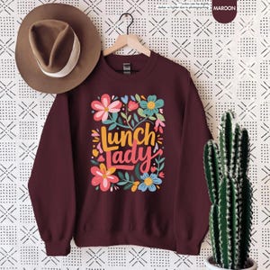 Pode incluir: Camisola bord&ocirc; com o texto "Lunch Lady" em um design floral colorido. O design inclui flores rosa, azuis e amarelas com folhas verdes. Um chap&eacute;u marrom est&aacute; pendurado acima da camisola.