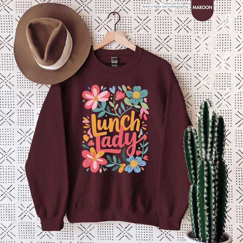 Pode incluir: Camisola bord&ocirc; com o texto "Lunch Lady" em um design floral colorido. O design inclui flores rosa, azuis e amarelas com folhas verdes. Um chap&eacute;u marrom est&aacute; pendurado acima da camisola.