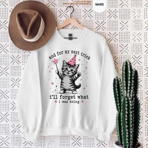 May include: A white long-sleeve sweatshirt featuring a graphic of a gray tabby kitten wearing a pink party hat and holding a star wand. The text reads, "and for my next trick i'll forget what i was doing," surrounded by small pink stars.