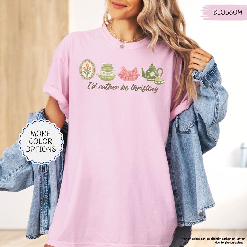 Pu&ograve; includere: T-shirt rosa chiaro con la scritta "I'd rather be thrifting" e una grafica di utensili da cucina vintage. La grafica include un design floreale incorniciato, ciotole, un piatto a forma di pollo e una teiera. La maglietta &egrave; color Blossom.