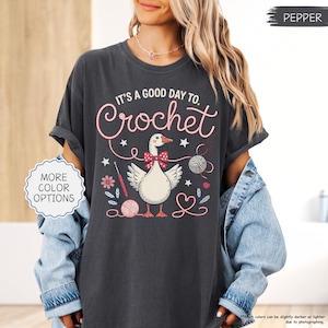 May include: Dark gray t-shirt with the text "IT'S A GOOD DAY TO. Crochet" in pink script. The shirt features a cartoon goose wearing a bow tie, surrounded by yarn balls, flowers, and crochet hooks. The shirt is a relaxed fit.