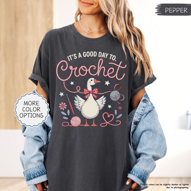 May include: Dark gray t-shirt with the text "IT'S A GOOD DAY TO. Crochet" in pink script. The shirt features a cartoon goose wearing a bow tie, surrounded by yarn balls, flowers, and crochet hooks. The shirt is a relaxed fit.