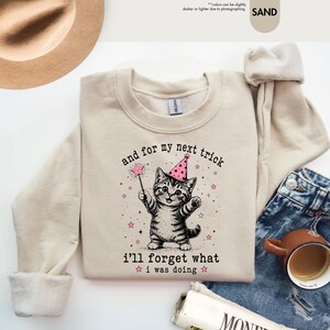 May include: A sand-colored crewneck sweatshirt featuring a graphic of a tabby cat wearing a pink party hat and holding a star-tipped wand. The text reads, "and for my next trick i'll forget what i was doing," surrounded by small pink stars.