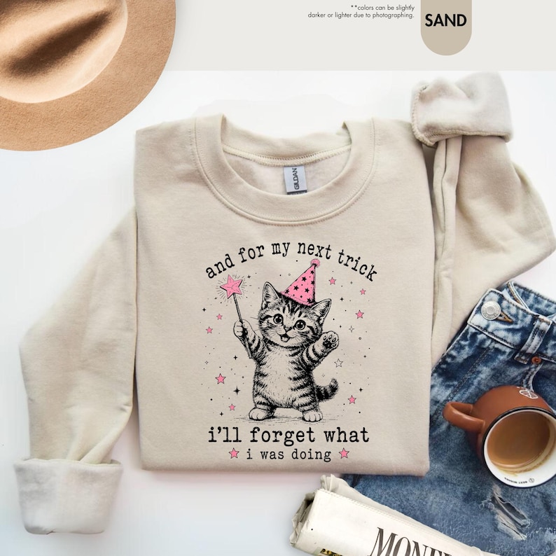 May include: A sand-colored crewneck sweatshirt featuring a graphic of a tabby cat wearing a pink party hat and holding a star-tipped wand. The text reads, "and for my next trick i'll forget what i was doing," surrounded by small pink stars.