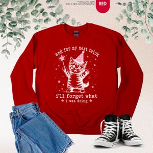 May include: A red long-sleeve sweatshirt featuring a white graphic of a kitten wearing a pink party hat and holding a magic wand. The text reads, and for my next trick i'll forget what i was doing, surrounded by small white stars.