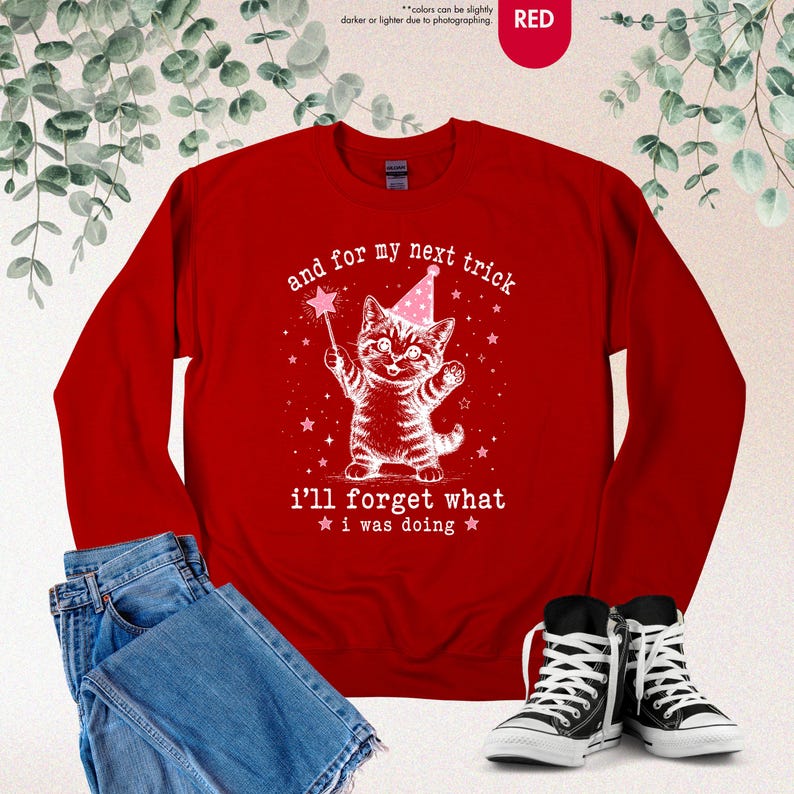 May include: A red long-sleeve sweatshirt featuring a white graphic of a kitten wearing a pink party hat and holding a magic wand. The text reads, and for my next trick i'll forget what i was doing, surrounded by small white stars.