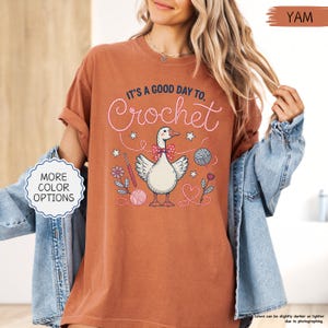 May include: A terracotta-colored t-shirt with the text "IT'S A GOOD DAY TO. Crochet" in white script. The graphic features a goose wearing a bow tie, surrounded by crochet-themed illustrations. The shirt has rolled-up sleeves and a relaxed fit.