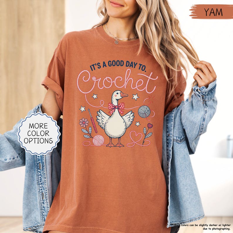 May include: A terracotta-colored t-shirt with the text "IT'S A GOOD DAY TO. Crochet" in white script. The graphic features a goose wearing a bow tie, surrounded by crochet-themed illustrations. The shirt has rolled-up sleeves and a relaxed fit.