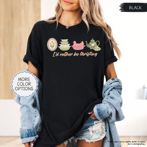Pu&ograve; includere: T-shirt nero con la scritta "I'd rather be thrifting" e una grafica di utensili da cucina vintage. La grafica include un design floreale incorniciato, ciotole impilate, un pollo rosa e una teiera. La maglietta ha le maniche arrotolate.