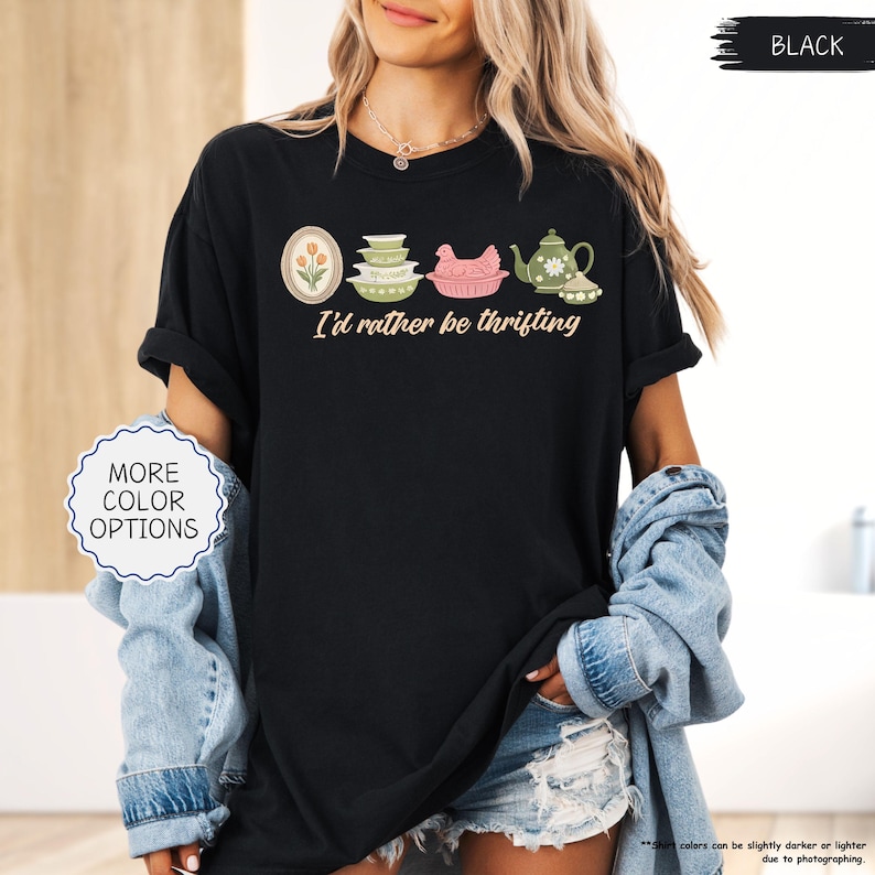 Pu&ograve; includere: T-shirt nero con la scritta "I'd rather be thrifting" e una grafica di utensili da cucina vintage. La grafica include un design floreale incorniciato, ciotole impilate, un pollo rosa e una teiera. La maglietta ha le maniche arrotolate.