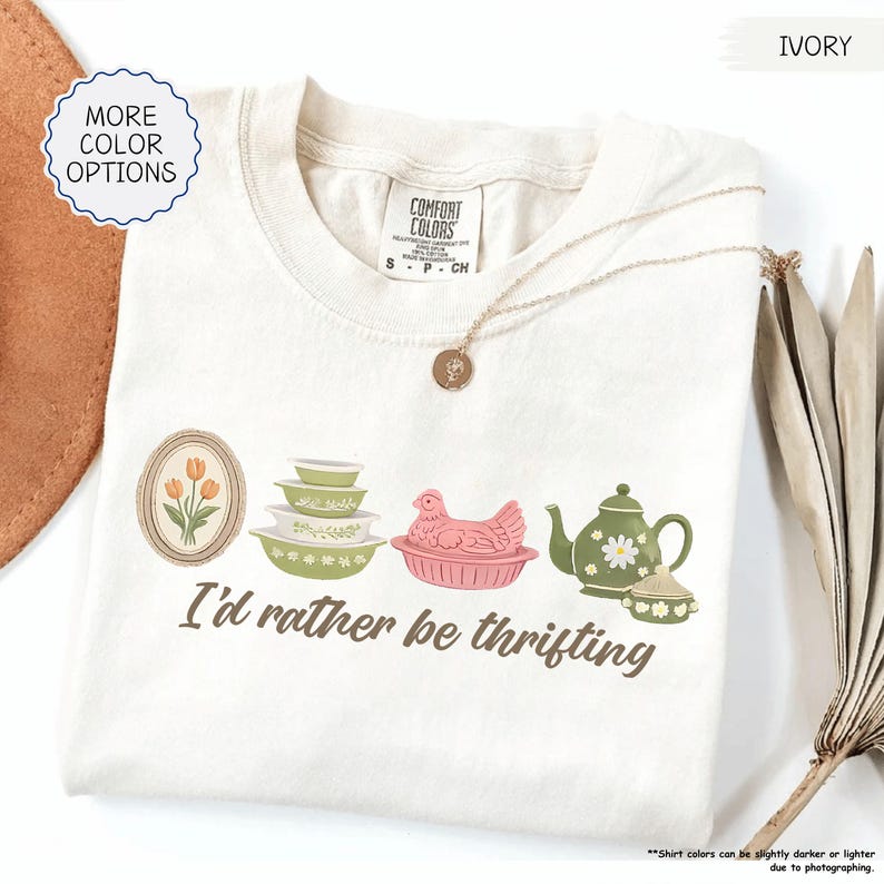 Pu&ograve; includere: T-shirt color avorio con la frase "I'd rather be thrifting" e illustrazioni in stile vintage: un fiore incorniciato, ciotole, una gallina, una teiera e una zuccheriera. La maglietta ha un girocollo e maniche corte.