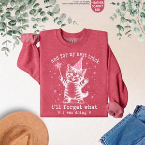 May include: A heather scarlet red crewneck sweatshirt featuring a white graphic of a kitten wearing a party hat and holding a star wand. The text reads, "and for my next trick i'll forget what i was doing," surrounded by small stars.
