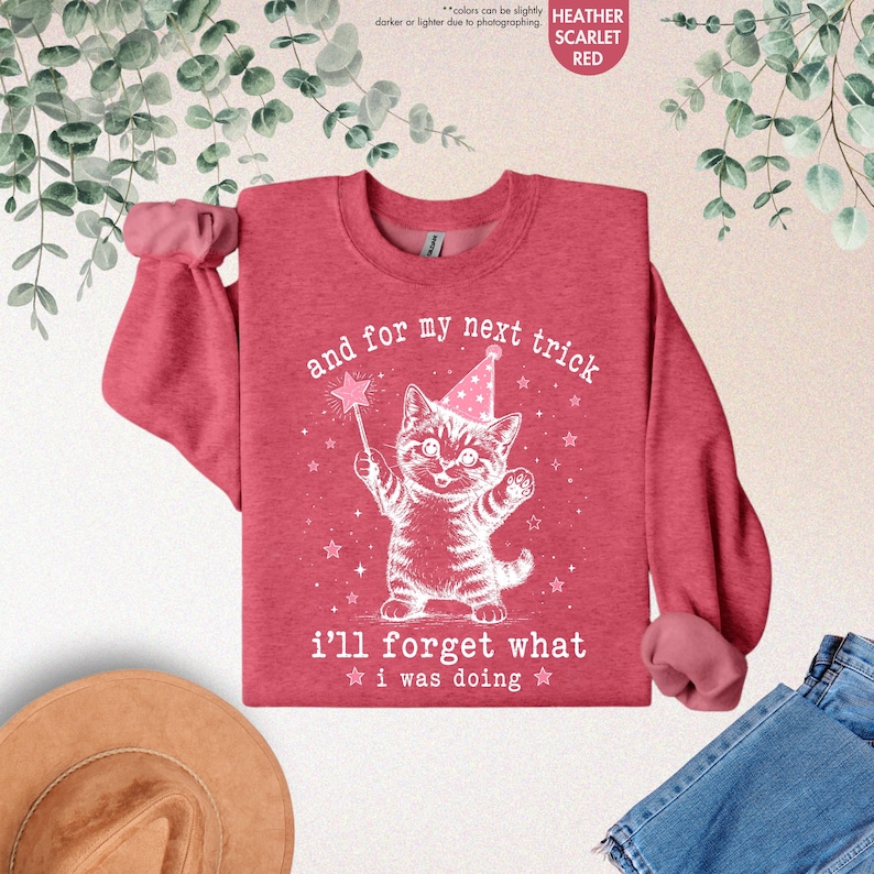 May include: A heather scarlet red crewneck sweatshirt featuring a white graphic of a kitten wearing a party hat and holding a star wand. The text reads, "and for my next trick i'll forget what i was doing," surrounded by small stars.