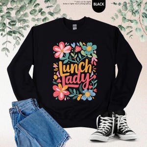 Pode incluir: Camisola preta com a frase "Lunch Lady" num design floral colorido. O design inclui flores rosa, azuis e amarelas com folhas verdes. A camisola &eacute; mostrada com cal&ccedil;as de ganga azuis e t&eacute;nis pretos e brancos.