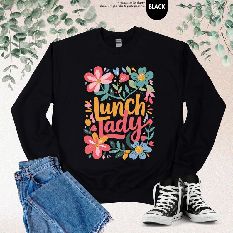 Pode incluir: Camisola preta com a frase "Lunch Lady" num design floral colorido. O design inclui flores rosa, azuis e amarelas com folhas verdes. A camisola &eacute; mostrada com cal&ccedil;as de ganga azuis e t&eacute;nis pretos e brancos.