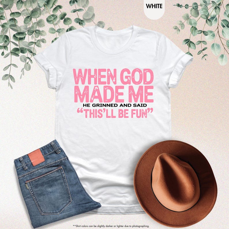 May include: White t-shirt with the text "WHEN GOD MADE ME HE GRINNED AND SAID 'THIS'LL BE FUN'" in pink. The image also shows blue jeans and a brown hat. The shirt is a casual wear item.