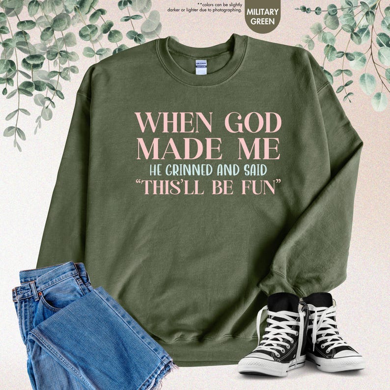 May include: Military green sweatshirt with the text "WHEN GOD MADE ME HE GRINNED AND SAID 'THIS'LL BE FUN'" in pink lettering. The sweatshirt is paired with blue jeans and black and white sneakers.