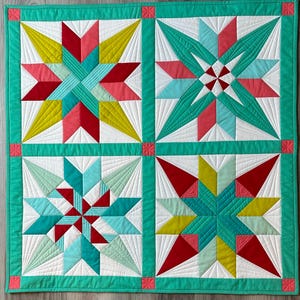 May include: A quilt featuring four distinct star patterns in shades of red, yellow, teal, and white. The quilt is bordered with a teal trim and is displayed on a teal surface.
